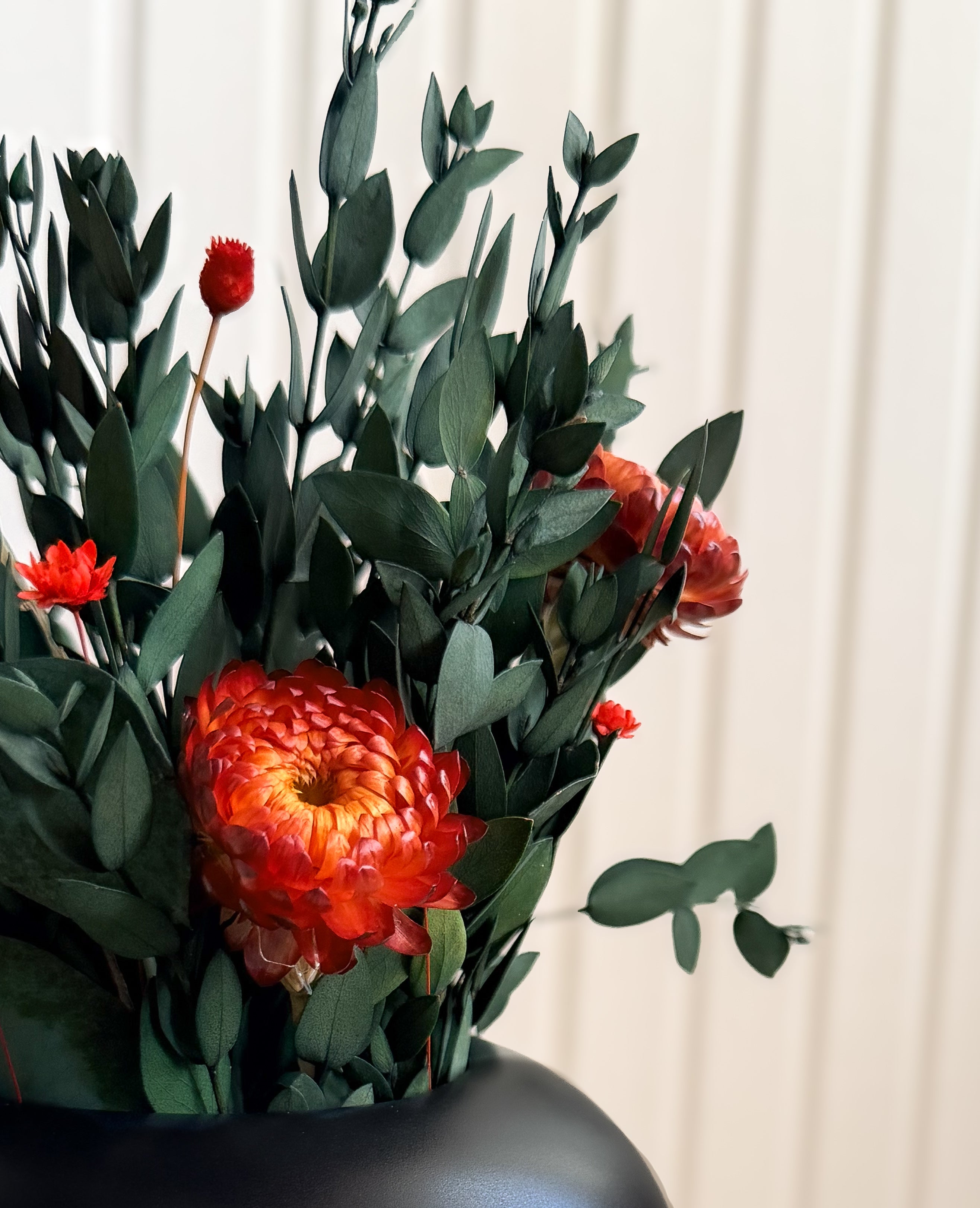 Närbild av Gåvoset 'KVÄLLSTID' arrangerat i svart Vas 'VALV' med mörkgrönt bladverk och orange blommor.